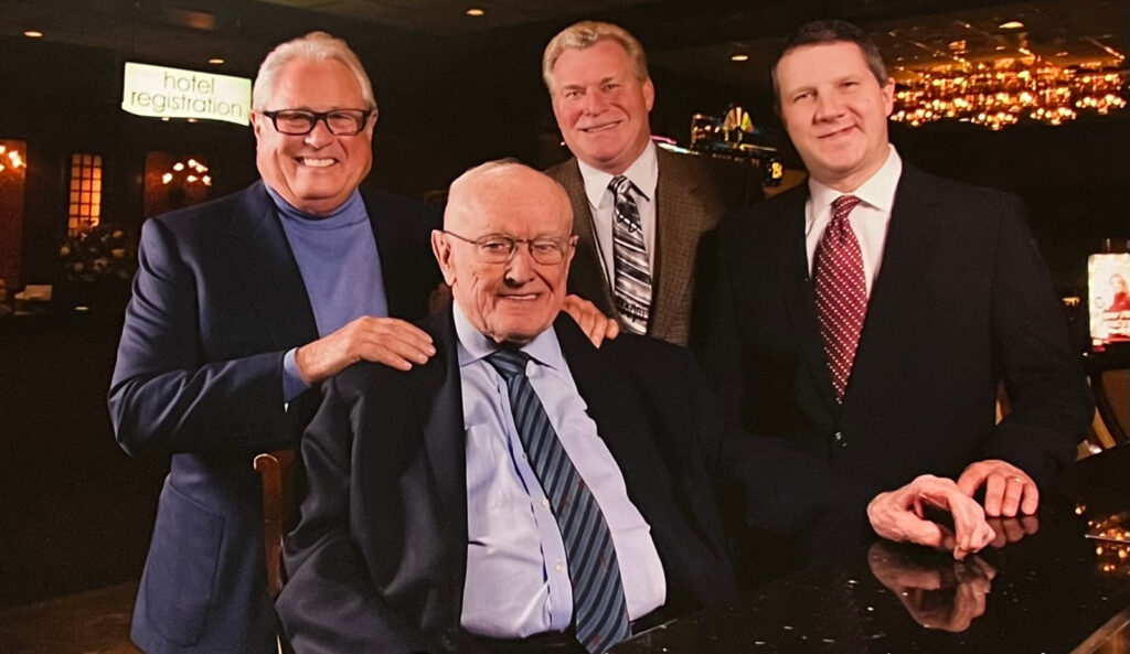 Four men in suits are smiling; one is seated while the others stand around him, with a hotel registration sign subtly hinting at history in the background.